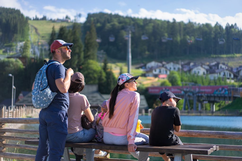 A family enjoys a sunny day sitting on a bench in Bukovel, Ukraine, with scenic mountain views.